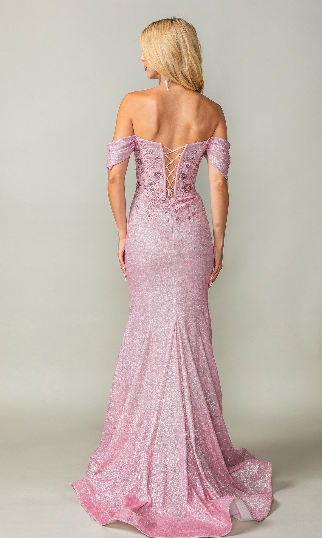 A woman models the Dancing Queen 4362, an off-shoulder long prom dress in shimmery pink with a lace-up back and floral embellishments, standing with her back to the camera.